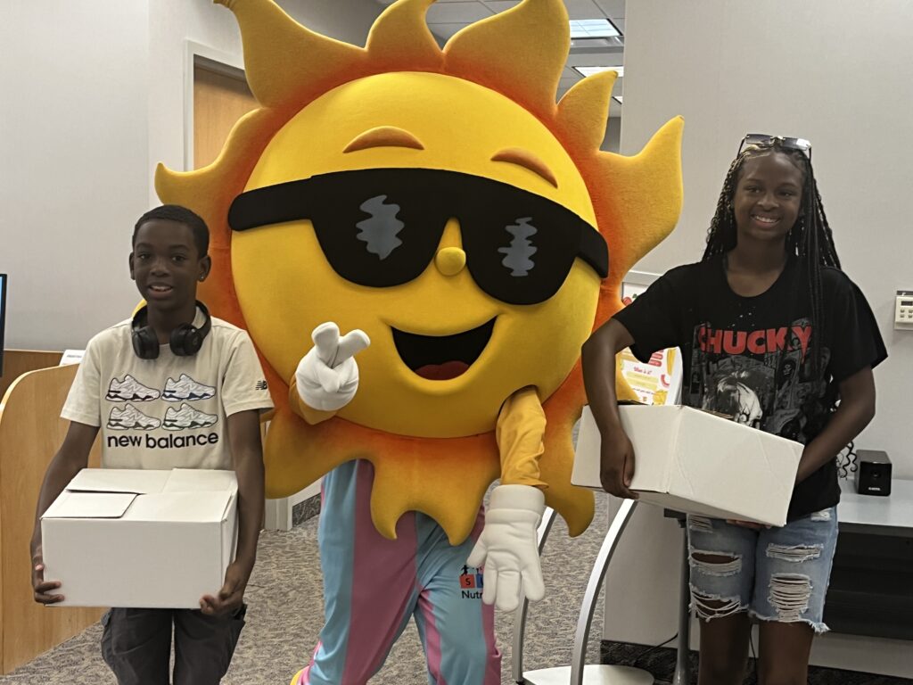 two children holding boxes of meals to-go with a large sun mascot, Ray F. Sunshine